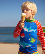 Child standing on the beach with the ocean in the background and holding a i cream. Wearing a blue rash vest with red and white stripes 