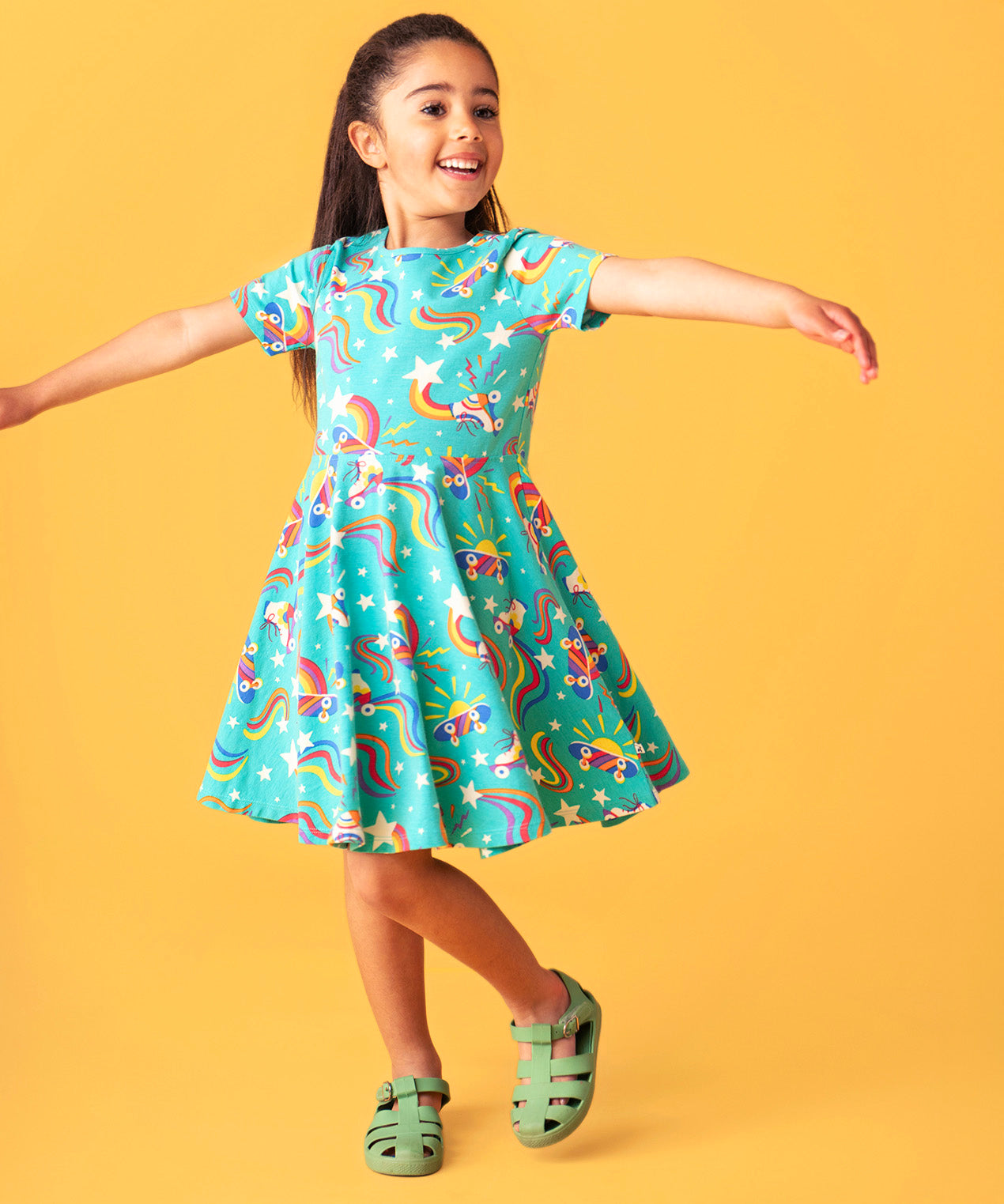 Child smiling and dancing in front of a light orange background. Wearing the turquoise skater dress with repeating roller blades and skateboard prints.