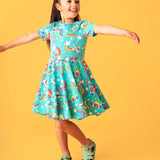 Child smiling and dancing in front of a light orange background. Wearing the turquoise skater dress with repeating roller blades and skateboard prints.