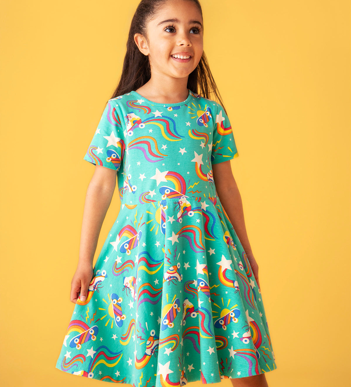 Child smiling and standing in front of a light orange background. Wearing the turquoise skater dress with repeating roller blades and skateboard prints.