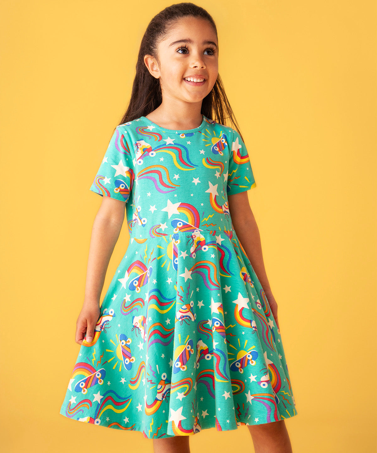 Child smiling and standing in front of a light orange background. Wearing the turquoise skater dress with repeating roller blades and skateboard prints.