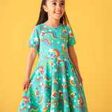 Child smiling and standing in front of a light orange background. Wearing the turquoise skater dress with repeating roller blades and skateboard prints.