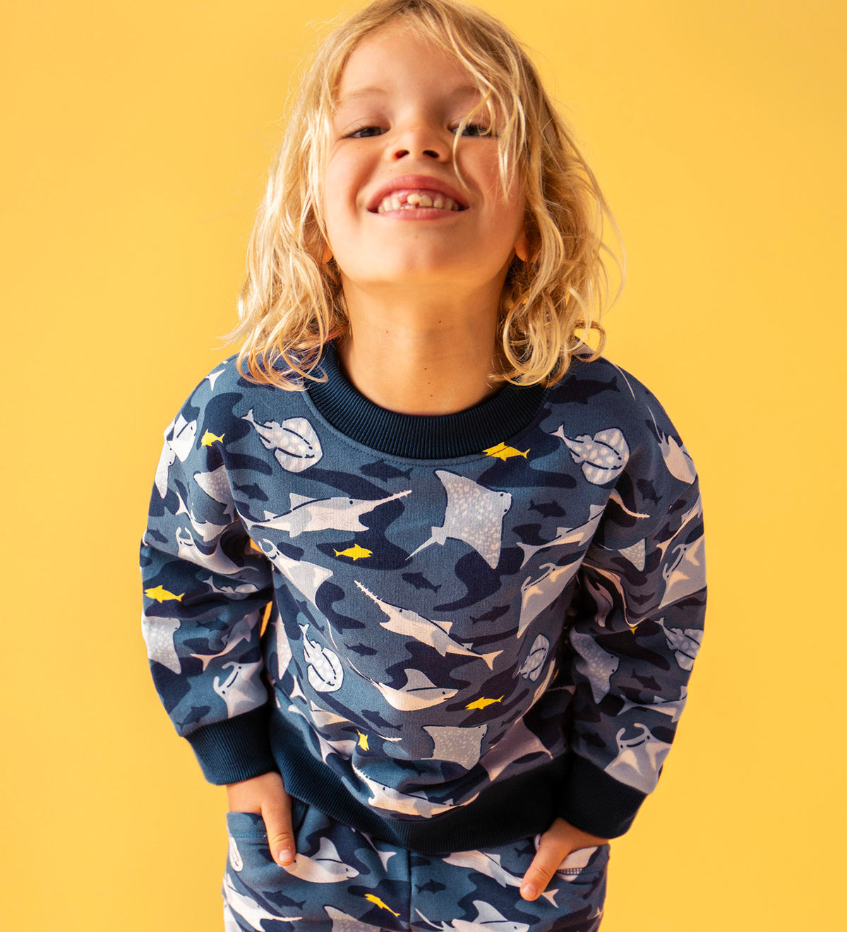 Child smiling and standing in front of a orange background. Wearing a dark blue sweatshirt and navy trim with a repeating manta ray print