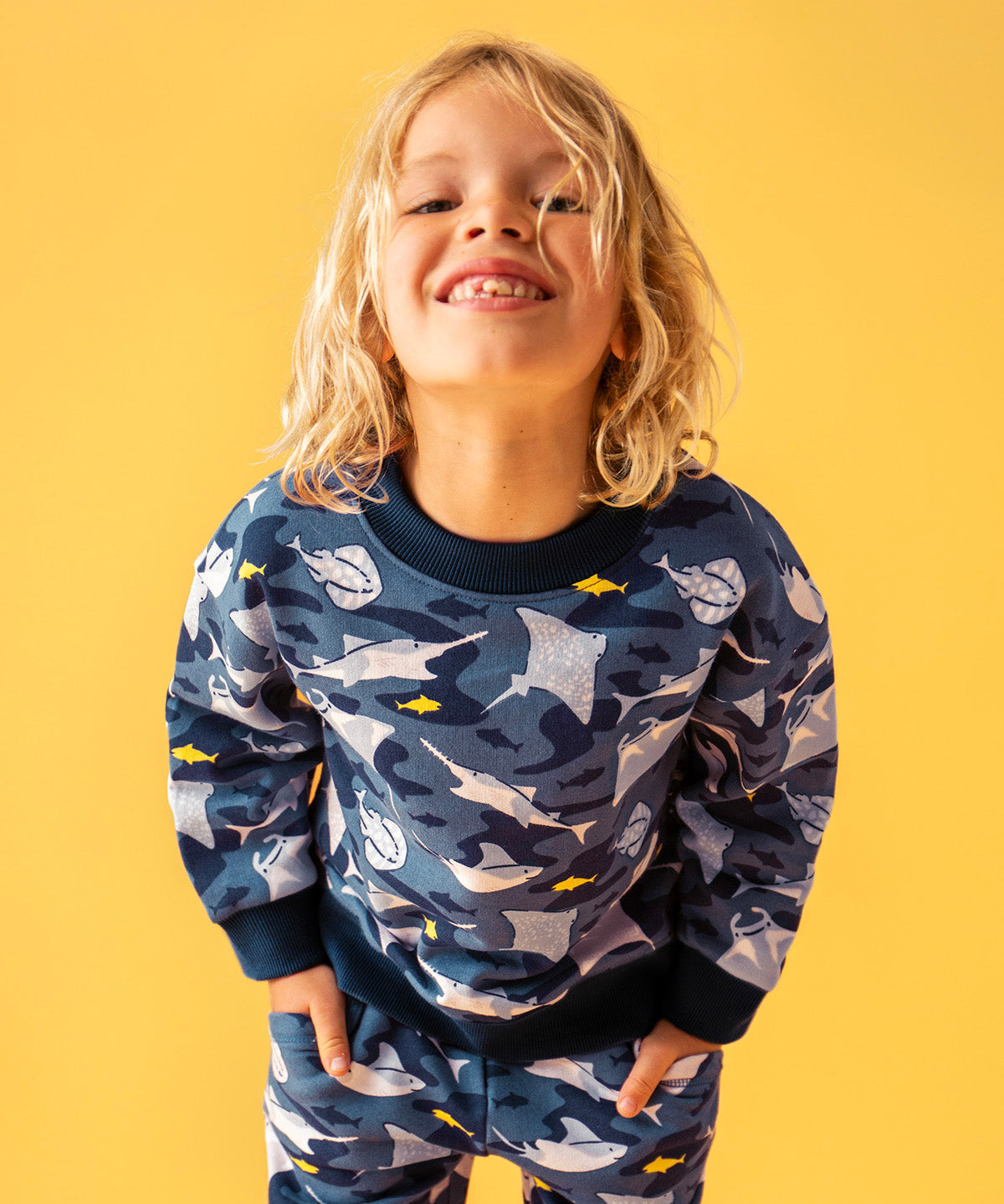 Child smiling and standing in front of a orange background. Wearing a dark blue sweatshirt and navy trim with a repeating manta ray print