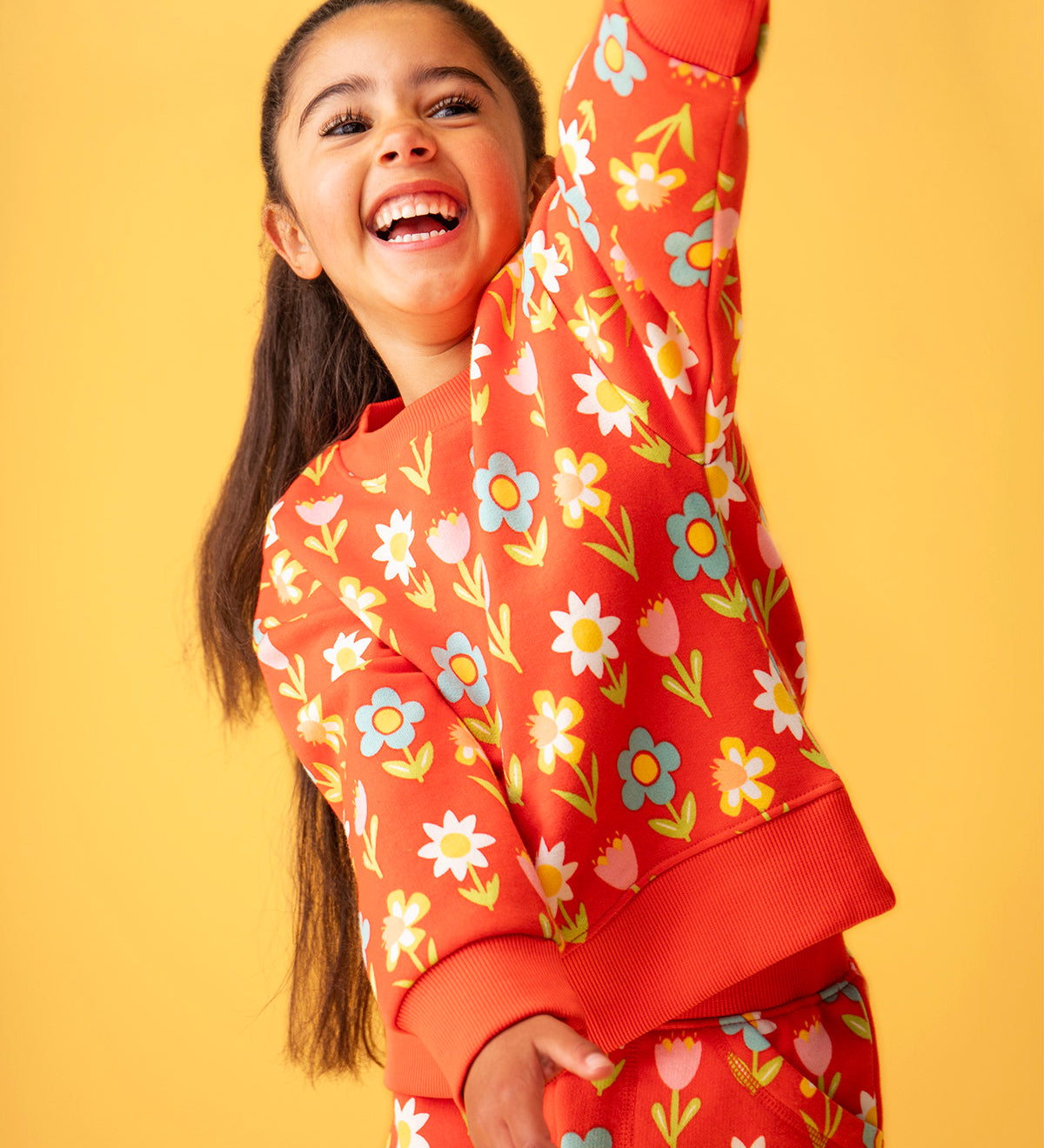 Toddler standing in front of a light orange background wearing the Frugi red sweatshirt with repeating pastel flower print and matching joggers