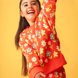 Toddler standing in front of a light orange background wearing the Frugi red sweatshirt with repeating pastel flower print and matching joggers