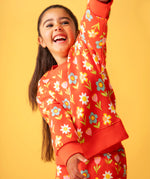 Toddler standing in front of a light orange background wearing the Frugi red sweatshirt with repeating pastel flower print and matching joggers