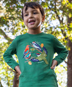 A child standing outside by trees and wearing the Frugi kids terrific applique top in holly green/birds. A dark green long sleeved GOTS organic top with different bird appliques