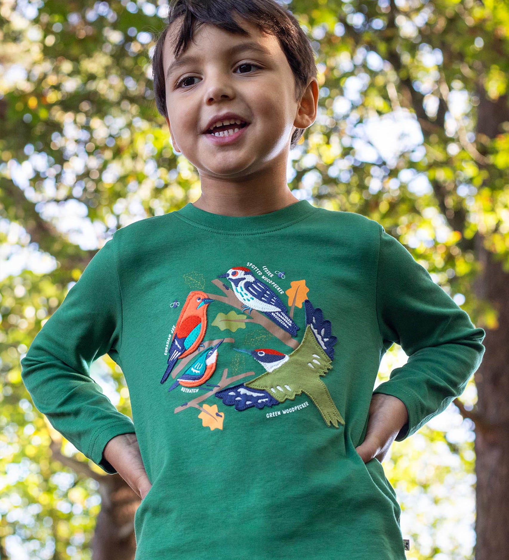 A child standing outside by trees and wearing the Frugi kids terrific applique top in holly green/birds. A dark green long sleeved GOTS organic top with different bird appliques