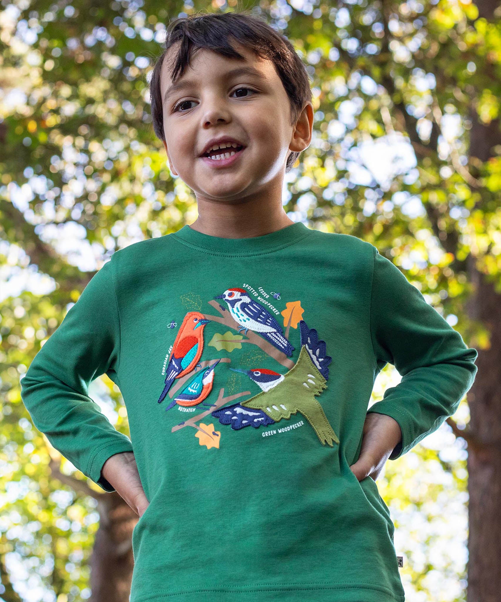 A child standing outside by trees and wearing the Frugi kids terrific applique top in holly green/birds. A dark green long sleeved GOTS organic top with different bird appliques