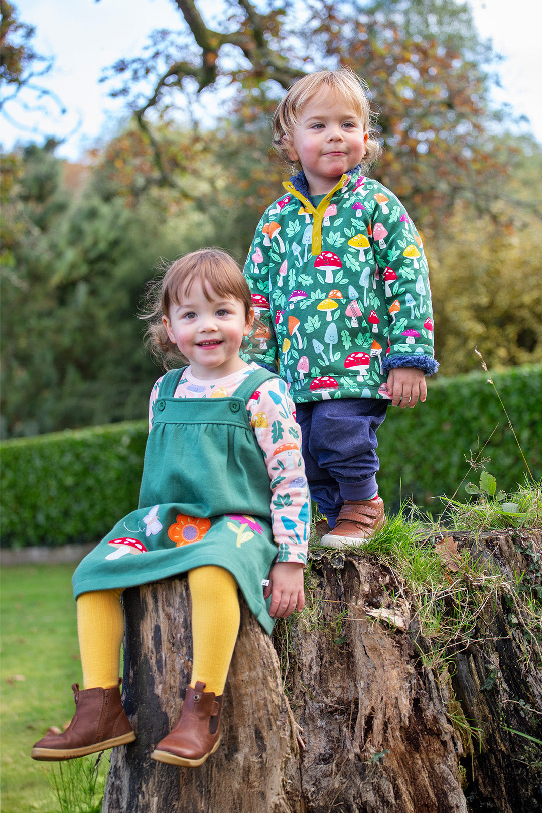 Two young children in Frugi Autumn Winter clothing from Babipur – green pinafore dress with floral appliqué and yellow tights, and green woodland print jacket – sitting and standing on a tree stump outdoors.