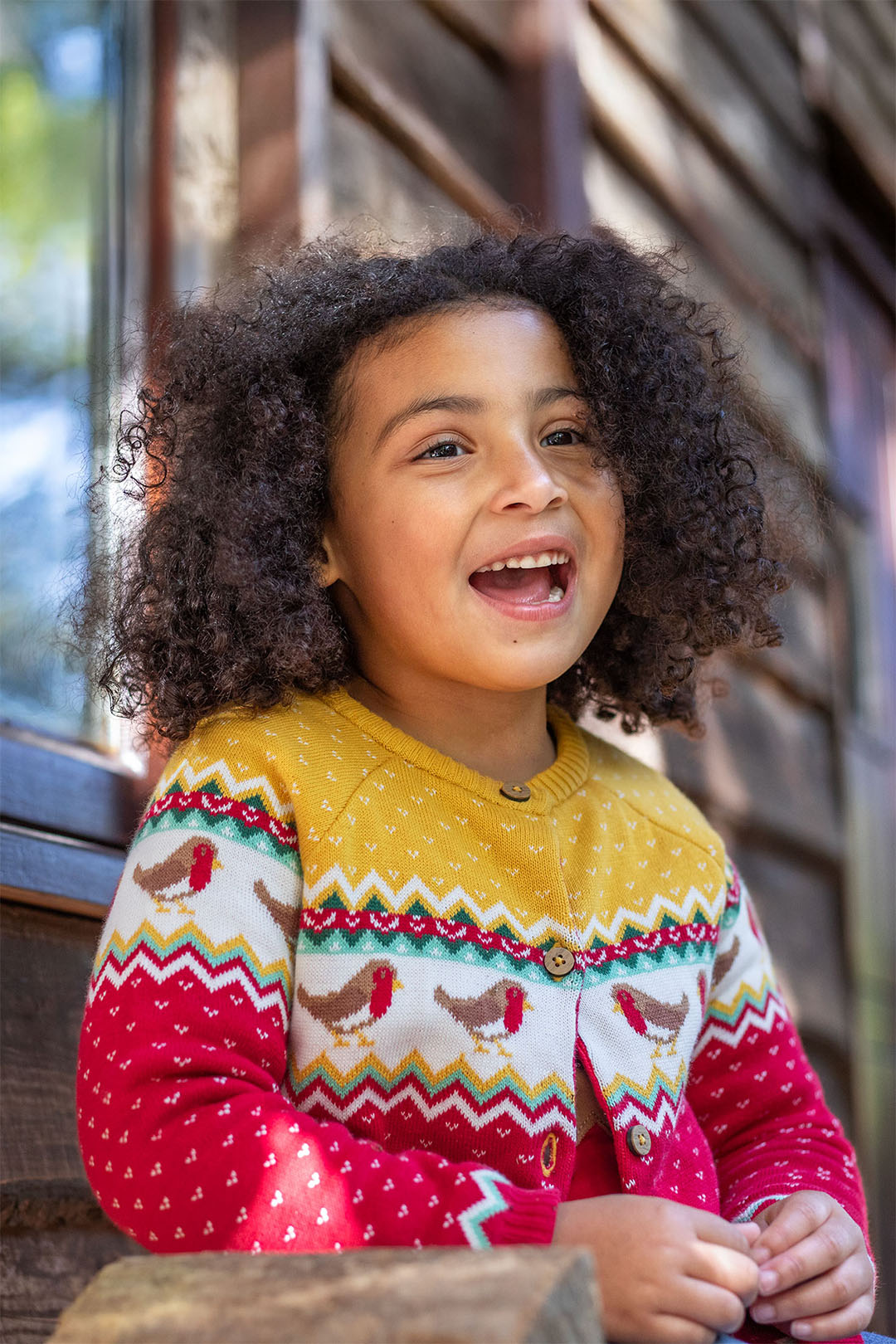 Smiley girl wearing a frugi fairisle kitted cardigan in red and yellow with robins by Frugi, organic cotton clothing for kids at Babipur - this image is to represent the brand at Babipur 