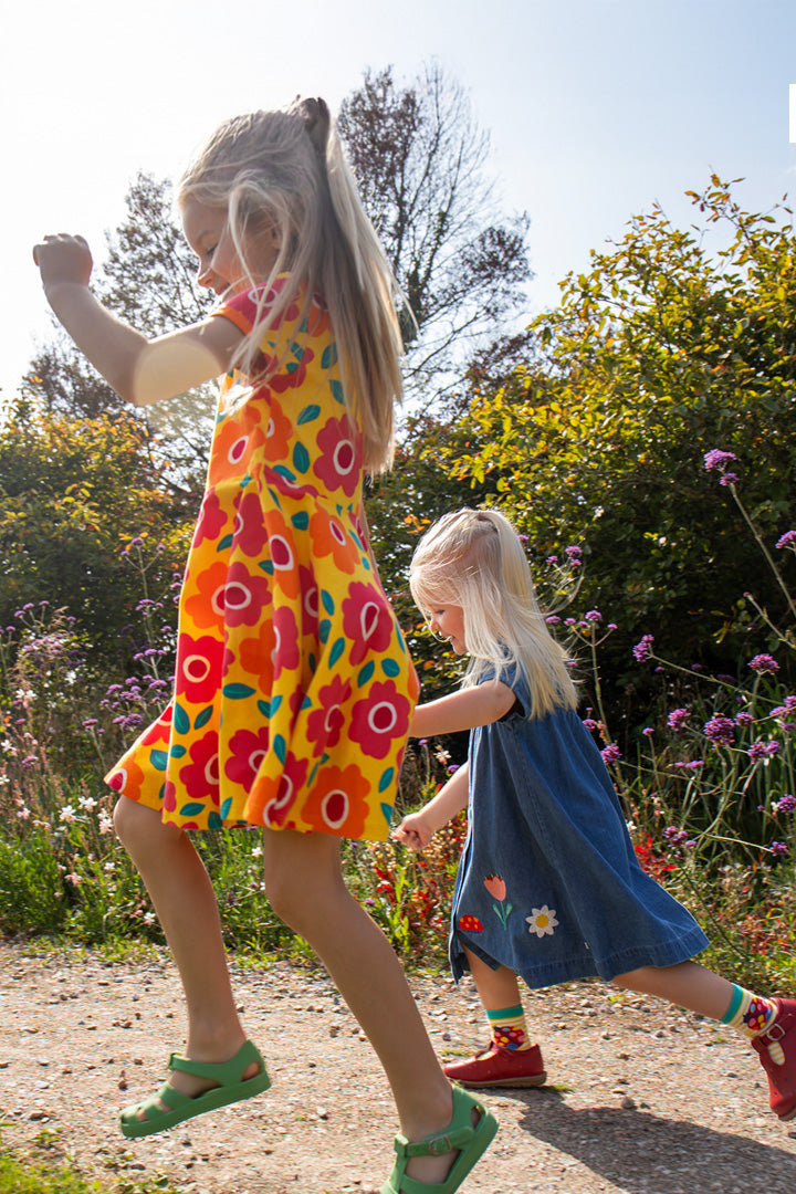 Two girls running in a field wearing Spring Frugi clothes from Babipur - yellow and pink floral organic cotton skater dress and rosemary denim dress with applique flowers around the hem. 