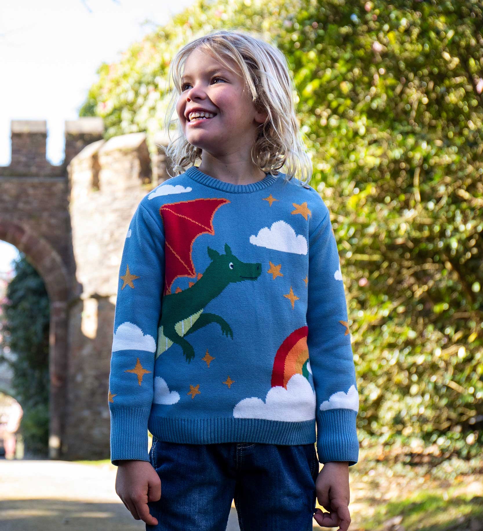 A child wearing the Frugi elwood dragon skies knitted jumper. A GOTS organic cotton jumper in light blue with a playful flying dragon design wrapping around the jumper. Features button fasteners on the shoulder. This children's knitted jumper is apart of a wide range of organic cotton clothing for children available here at Babipur. 