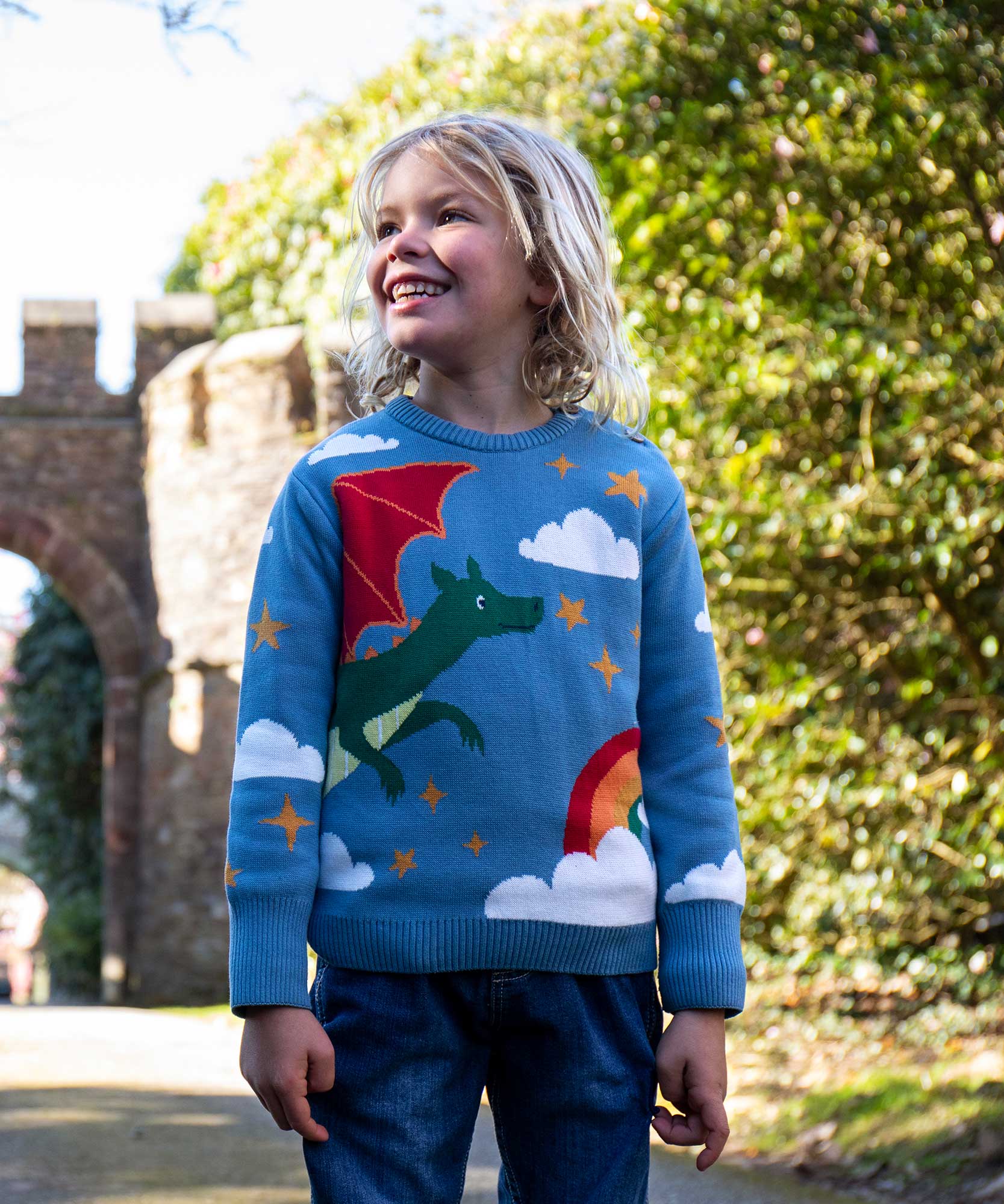 A child wearing the Frugi elwood dragon skies knitted jumper. A GOTS organic cotton jumper in light blue with a playful flying dragon design wrapping around the jumper. Features button fasteners on the shoulder. This children's knitted jumper is apart of a wide range of organic cotton clothing for children available here at Babipur. 
