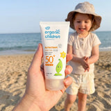 Adult hand holding a 150ml tube of Green People organic children SPF50 scent free sun cream showing scale with a child in the background