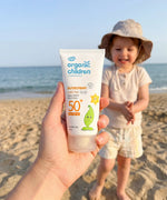 Adult hand holding a 150ml tube of Green People organic children SPF50 scent free sun cream showing scale with a child in the background