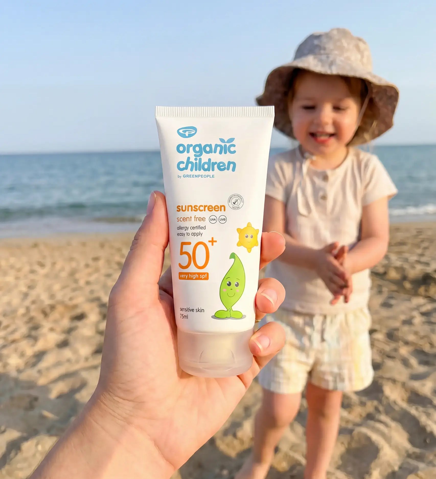 Adult hand holding a 150ml tube of Green People organic children SPF50 scent free sun cream showing scale with a child in the background