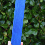A hand holding a Glückskäfer lerge blue rectangle wooden slat block against a blurred background of green foliage.