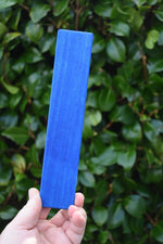 A hand holding a Glückskäfer lerge blue rectangle wooden slat block against a blurred background of green foliage.