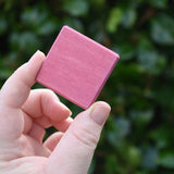 A hand holding a Glückskäfer small, square pink wooden slat block against a blurred background of green foliage.