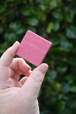 A hand holding a Glückskäfer small, square pink wooden slat block against a blurred background of green foliage.