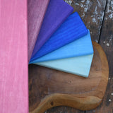 A fan of Glückskäfer rectangular wood slats in pink, mauve, purple, blue, and pale aqua, arranged on a rustic wooden board over weathered wood planks.