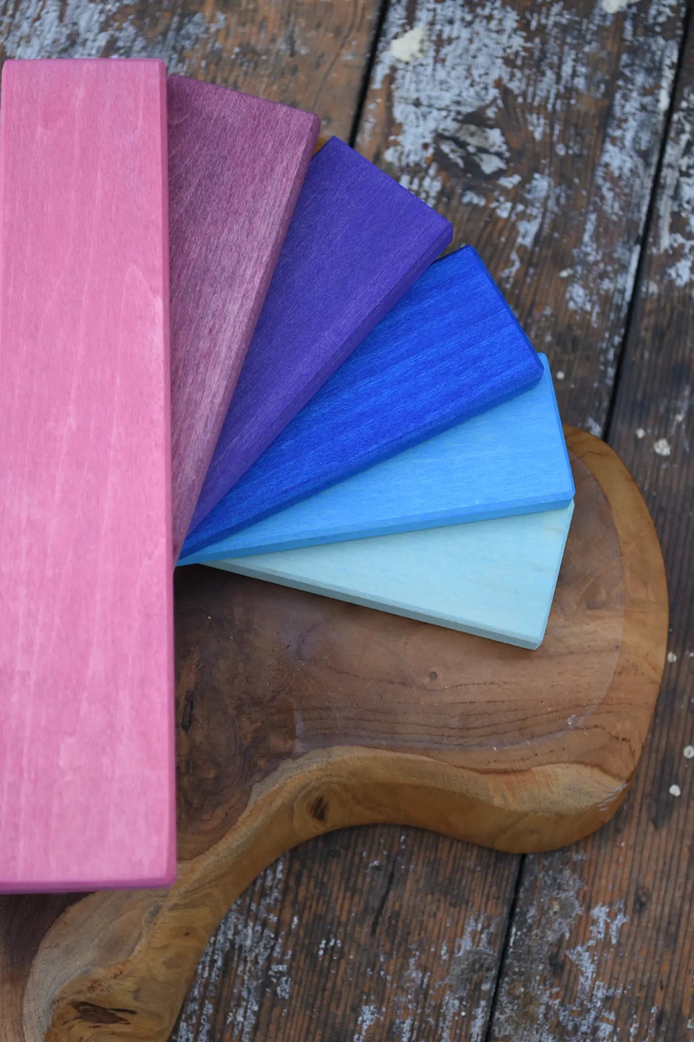 A fan of Glückskäfer rectangular wood slats in pink, mauve, purple, blue, and pale aqua, arranged on a rustic wooden board over weathered wood planks.