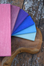 A fan of Glückskäfer rectangular wood slats in pink, mauve, purple, blue, and pale aqua, arranged on a rustic wooden board over weathered wood planks.