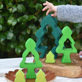 A child's hand is placing a dark green wooden tree-shaped piece from the Glückskäfer 6-Piece Fir Tree wooden tree stacking toy on a wooden board, next to other green and yellow wooden tree toys on a ceramic dish, with a leafy green garden background.