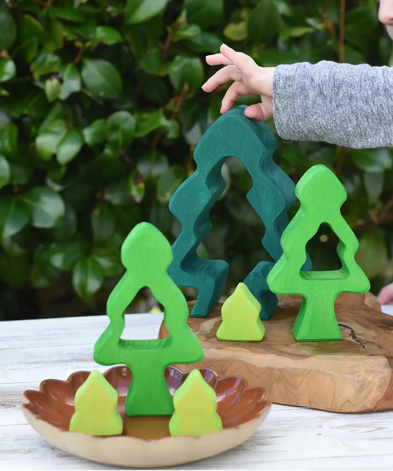 A child's hand is placing a dark green wooden tree-shaped piece from the Glückskäfer 6-Piece Fir Tree wooden tree stacking toy on a wooden board, next to other green and yellow wooden tree toys on a ceramic dish, with a leafy green garden background.