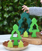 A child's hand is placing a dark green wooden tree-shaped piece from the Glückskäfer 6-Piece Fir Tree wooden tree stacking toy on a wooden board, next to other green and yellow wooden tree toys on a ceramic dish, with a leafy green garden background.