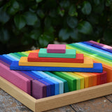 Glückskäfer colorful wooden slat blocks arranged in rainbow order, photographed outdoors on a rustic wooden surface with blurred green foliage in the background.