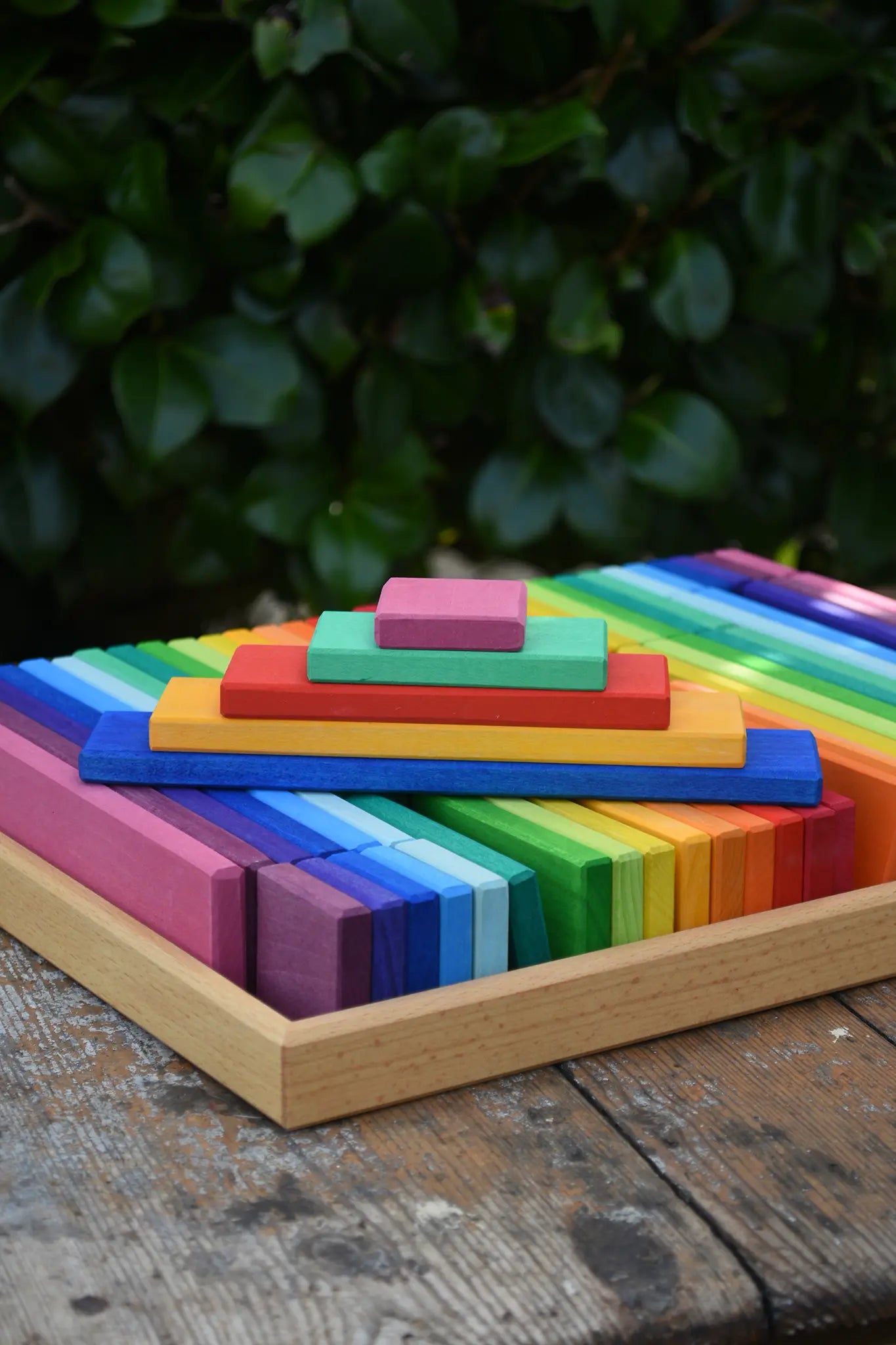 Glückskäfer colorful wooden slat blocks arranged in rainbow order, photographed outdoors on a rustic wooden surface with blurred green foliage in the background.