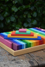 Glückskäfer colorful wooden slat blocks arranged in rainbow order, photographed outdoors on a rustic wooden surface with blurred green foliage in the background.