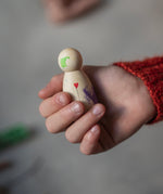 A child is holding a painted wooden Nin toy figure in their hand. The Nin is made from light wood with green and purple paint splotches, and a small red heart and a delicate black line coming from it, like a balloon. The Nin has been delicately painted by hand