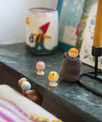 The colourful wooden fruit and vegetable figures are stood on a green/grey shelf with the blue one being held between a persons fingers and the orange figure in a sock, like a sock puppet