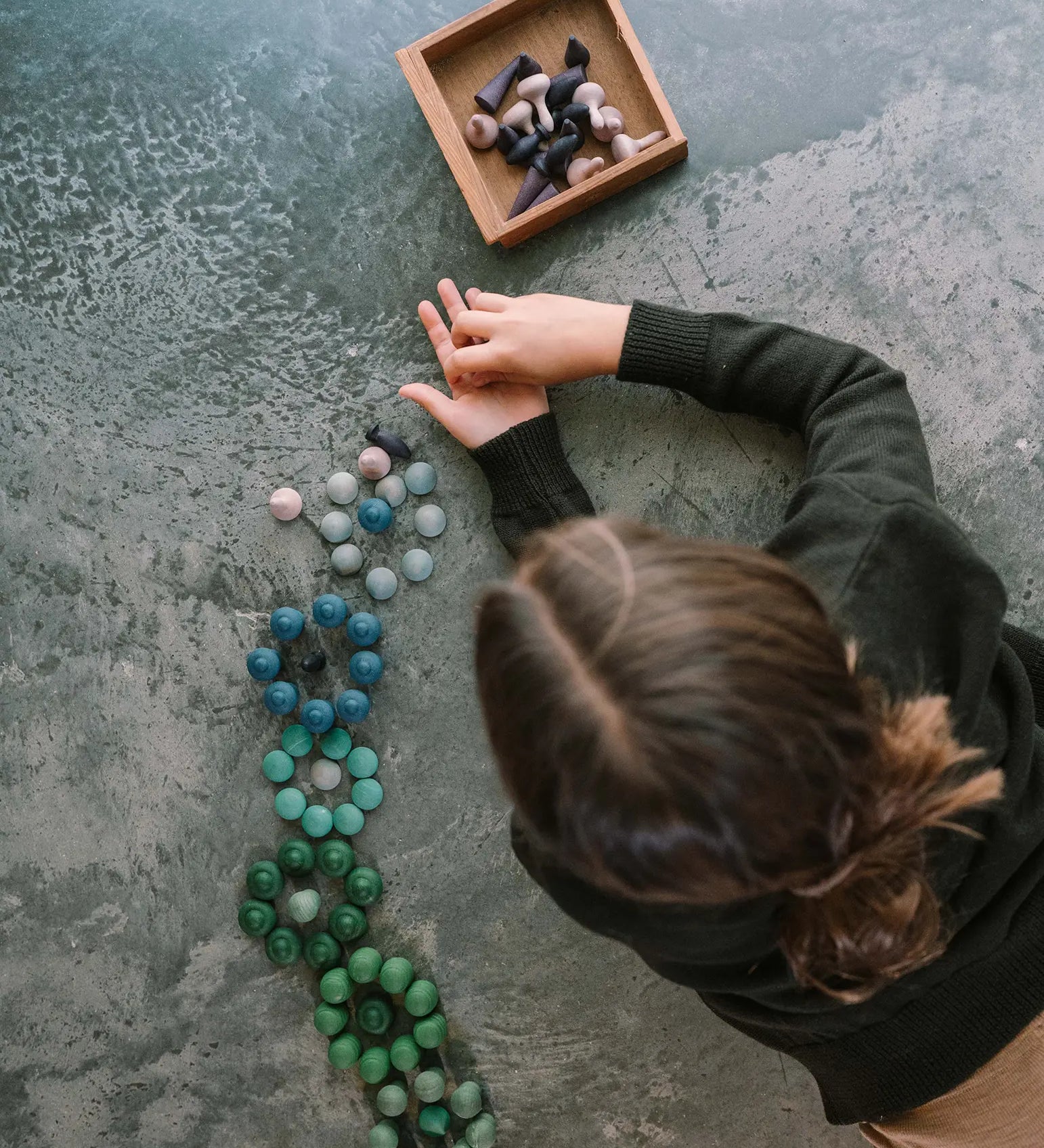 Child sorting Grapat's blue and green mixed handcrafted mandala pieces with a wooden box full of other purple mandala piece on a concrete floor