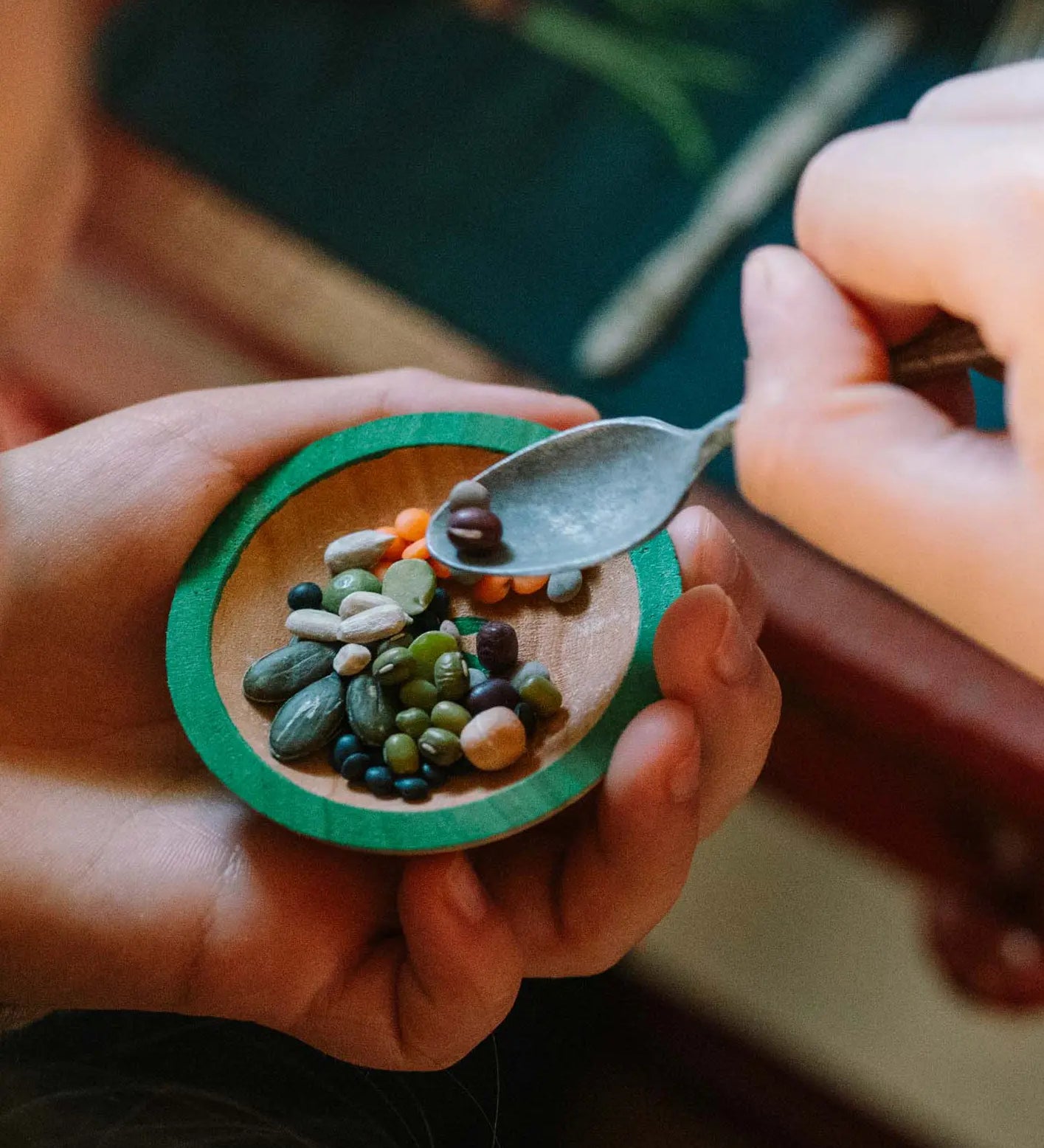Child's hand holding a toy dish by Grapat, made from wood with natural grain showing and green painted rim. The child is scooping seeds off the plate with a mini metal spoon.