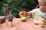 A child playing sat outside with the Grapat wooden yellow flowing bird next to two small purple plants on a garden table 
