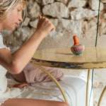 A child sitting on the bed next to a wooden table with the Grapat wooden orange wobbly bird resting on the table showing the hand painted purple and pink feather detailing