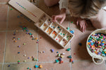 Child playing with the Grapat Mis Match & Bloom set, featuring a wooden sorting box, colourful geometric pieces and a woven basket on a terracotta-tiled floor.
