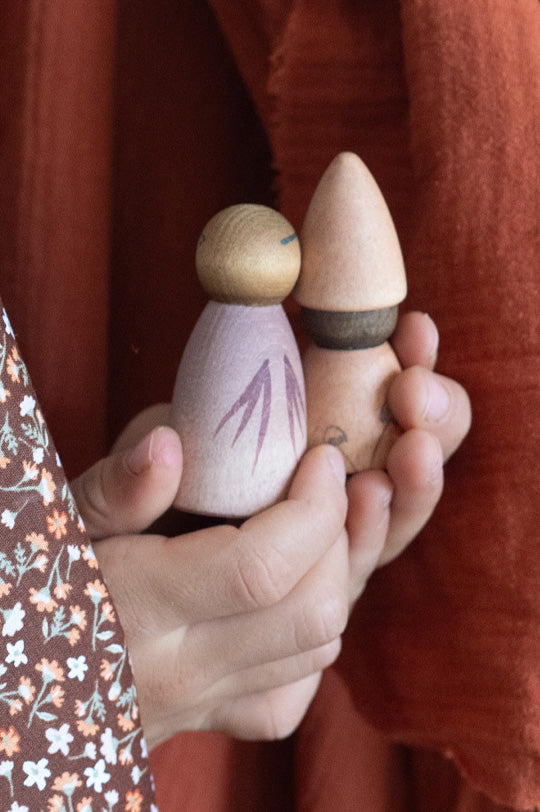 Child's hands holding two wooden peg dolls in purple and pink from Grapat's Moonlight Tale set, to represent Grapat Nins and Peg Dolls at Babipur.
