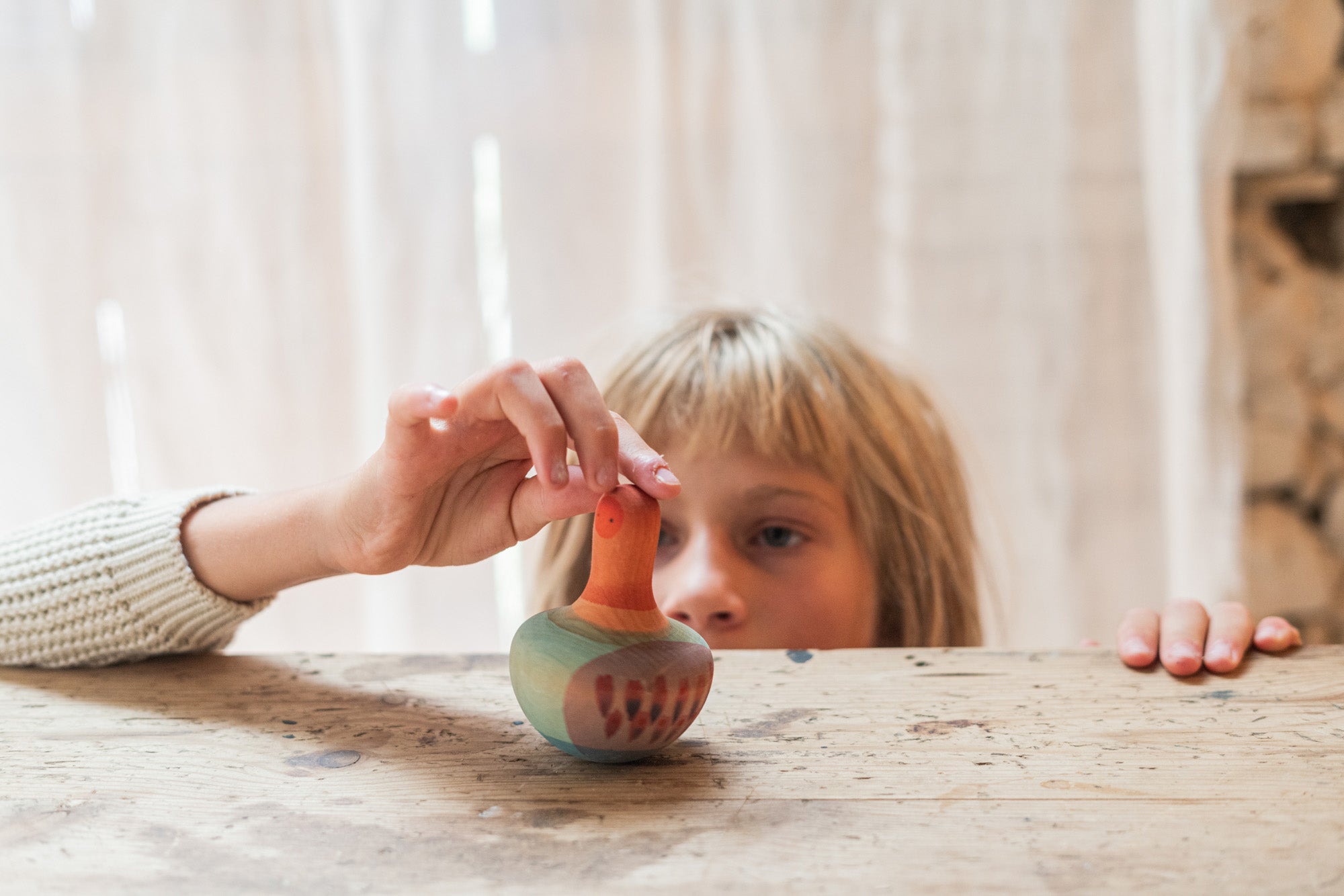 Child playing with the Grapat orange mellow wobbly bird. A close up showing the hand painted face with purple and pink feather detailing on a wooden table.