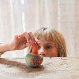 Child playing with the Grapat orange mellow wobbly bird. A close up showing the hand painted face with purple and pink feather detailing on a wooden table.