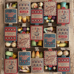 The Grapat Wooden Christmas Ornaments in their boxes, with the boxes lined up in three rows of 5. Each box has a whimsical Christmas design and the handmade and and painted ornaments can be used year after year.