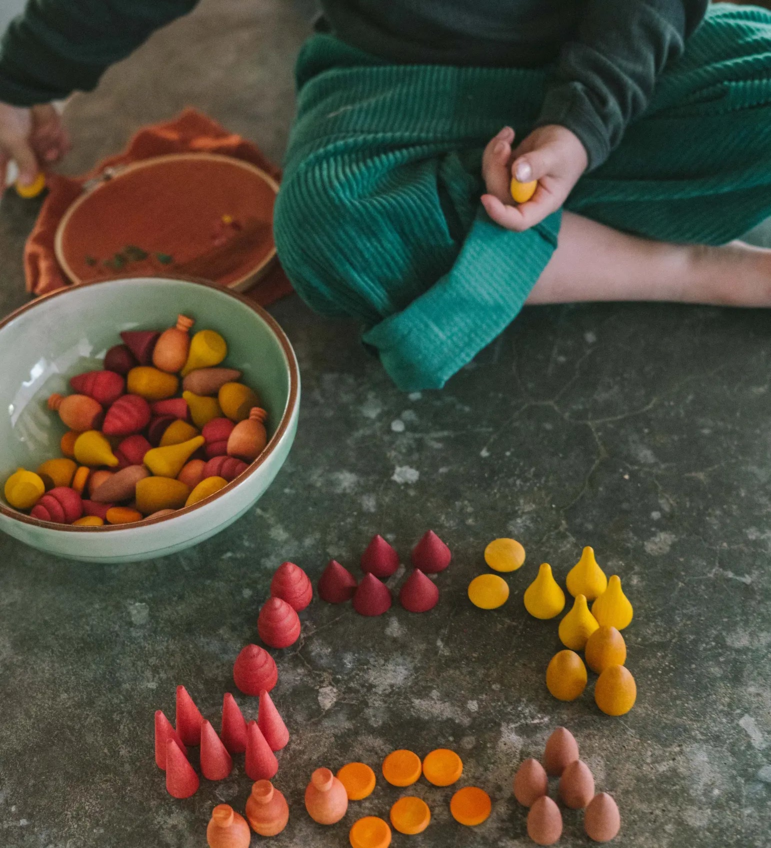 Grapat's handcrafted wooden mixed warm mandala pieces on a concrete surface standing next to a bowl full of other pieces and a sitting child