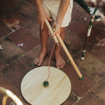 A child outside holding the Grapat wooden pendulum set showing the wooden pole dismantled on a stone floor next to a garden chair