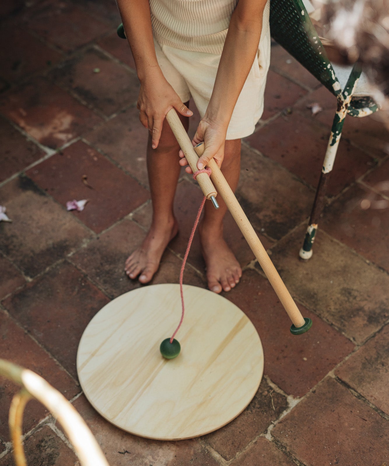 A child outside holding the Grapat wooden pendulum set showing the wooden pole dismantled on a stone floor next to a garden chair
