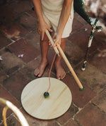 A child outside holding the Grapat wooden pendulum set showing the wooden pole dismantled on a stone floor next to a garden chair